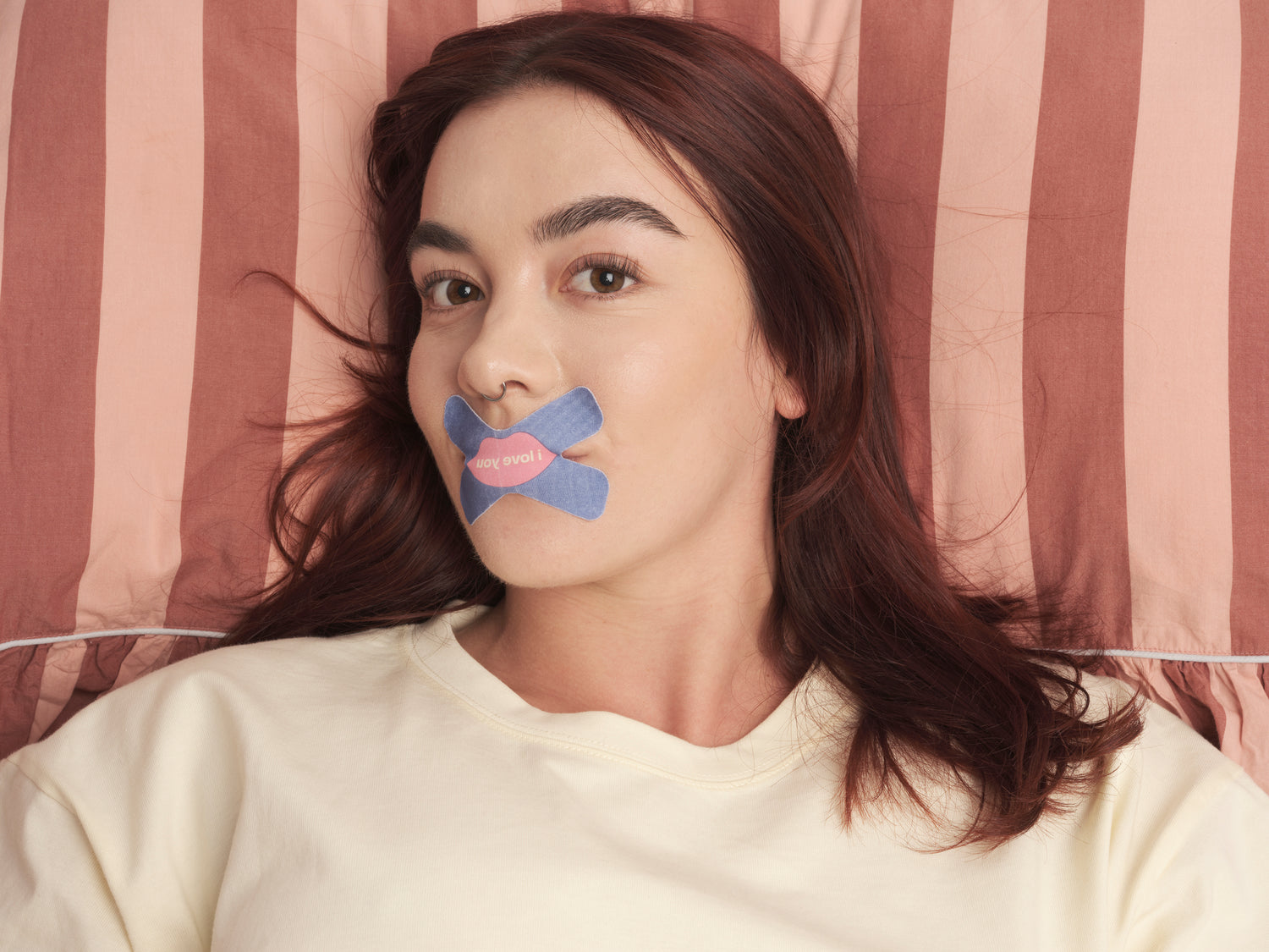 We shut it cross mouth tape on a female model laying in bed with striped duvet cover